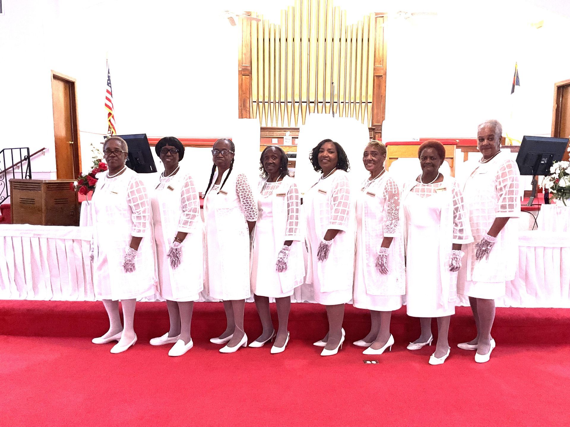 Group of women in white dresses and gloves standing on a red carpet in a church.