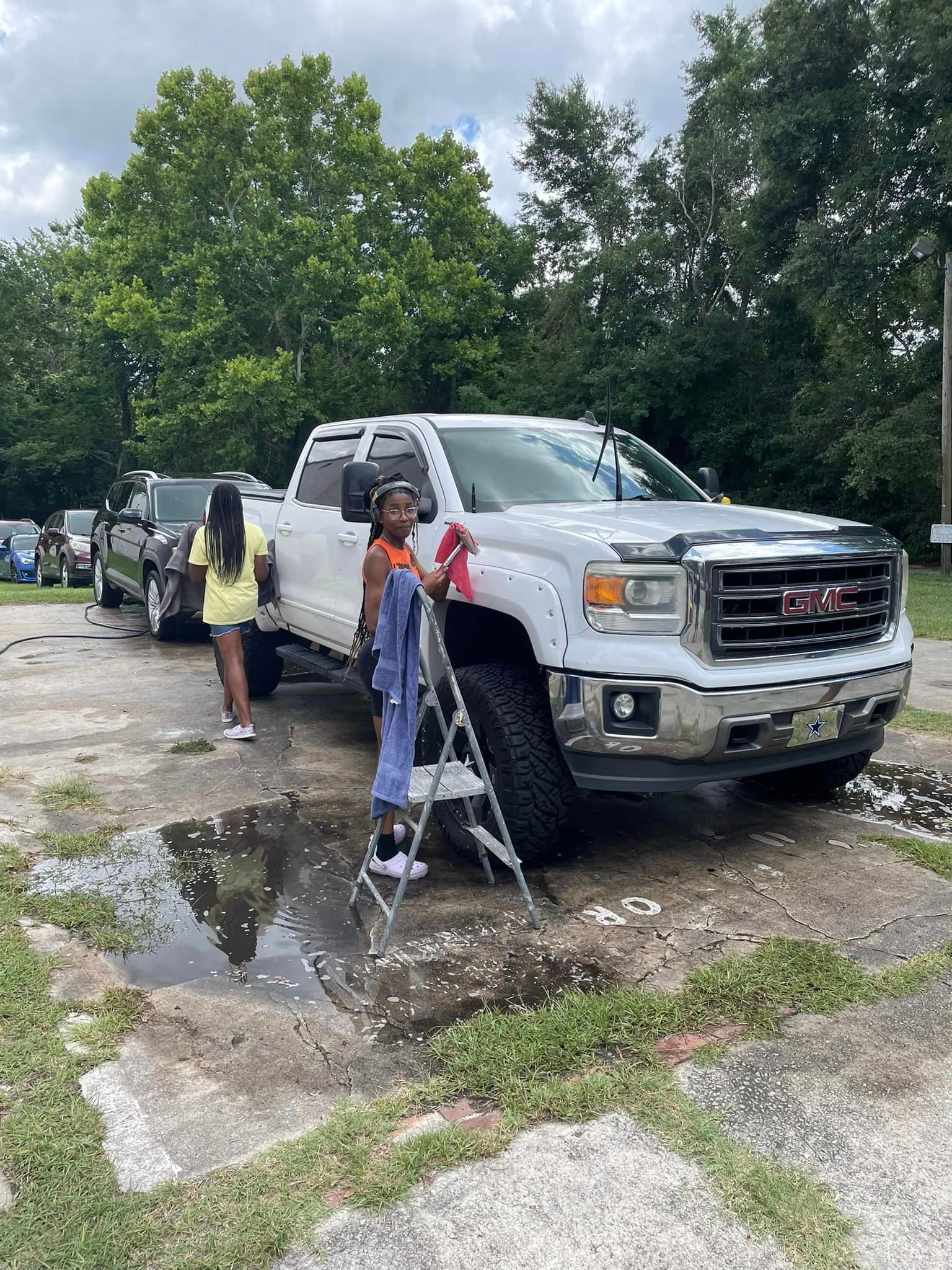 Two people washing a white GMC truck outdoors. One stands on a step stool.