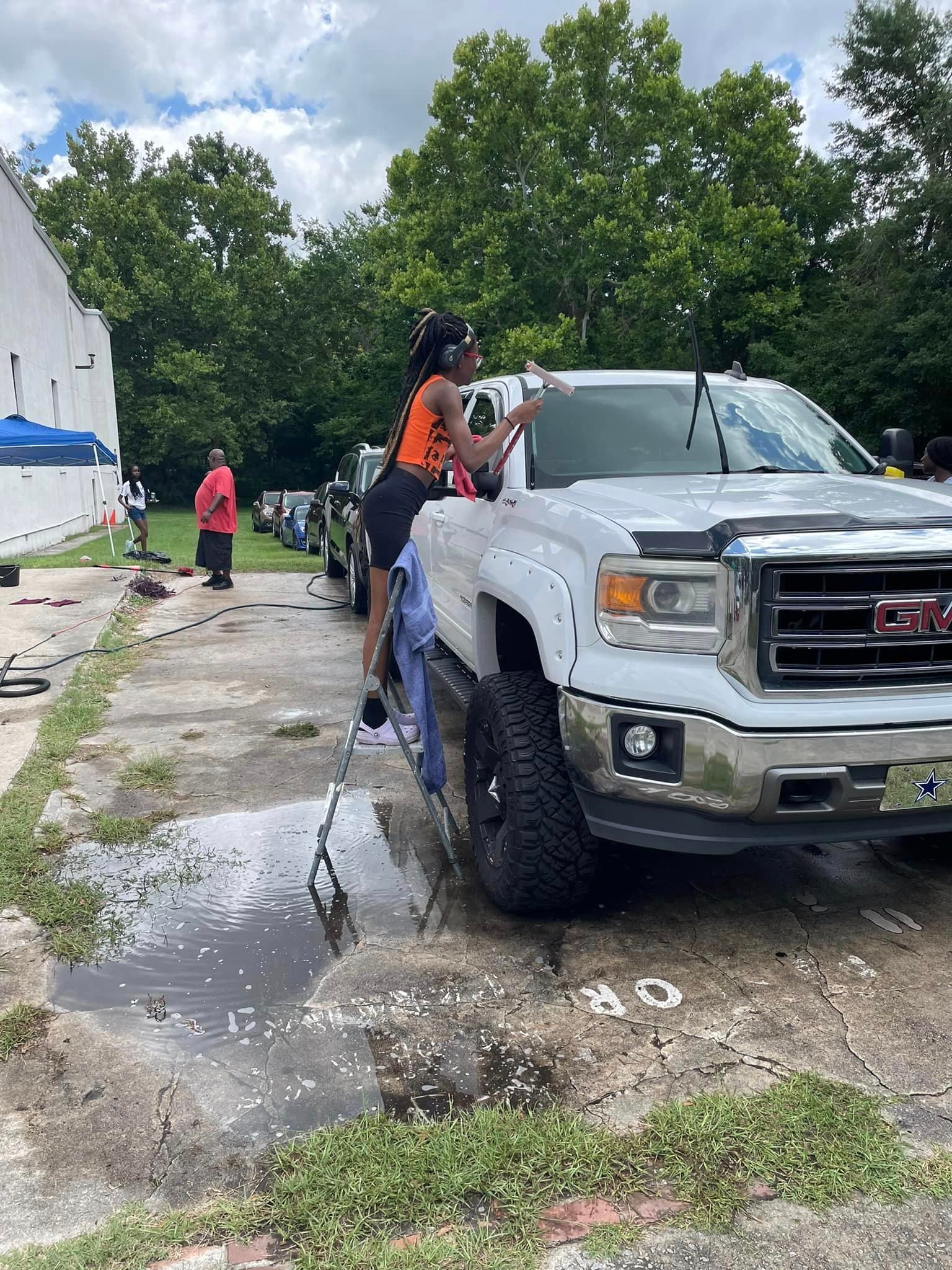 Woman washing a white GMC truck with a brush, standing on a step stool; outdoors.