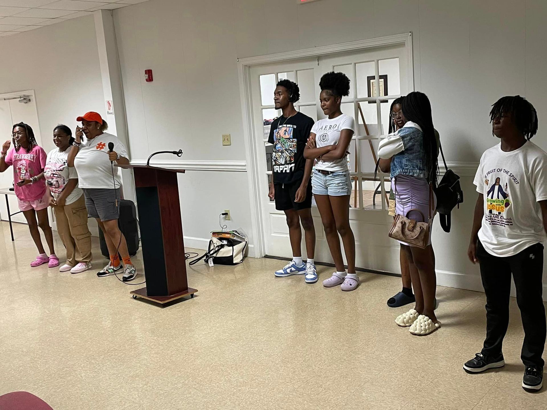 A group of people standing, some near a podium indoors. Several people appear to be listening.