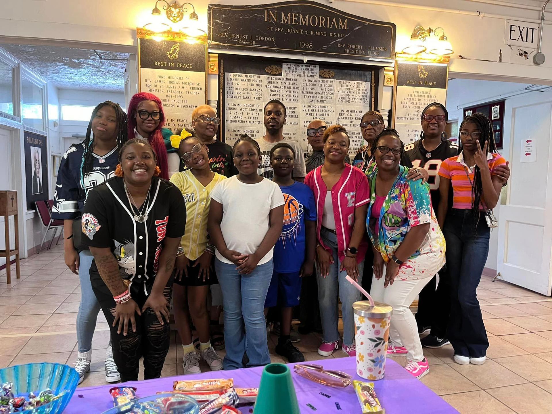 Group of people smiling, posing in front of a memorial board. Table with purple cloth, treats.