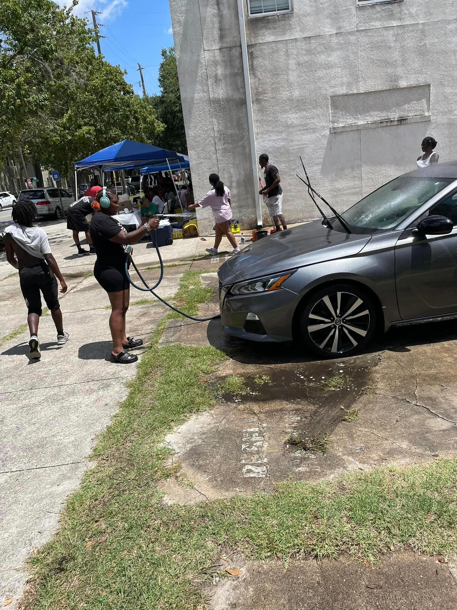 People washing a gray car outside a building; a car wash is underway.