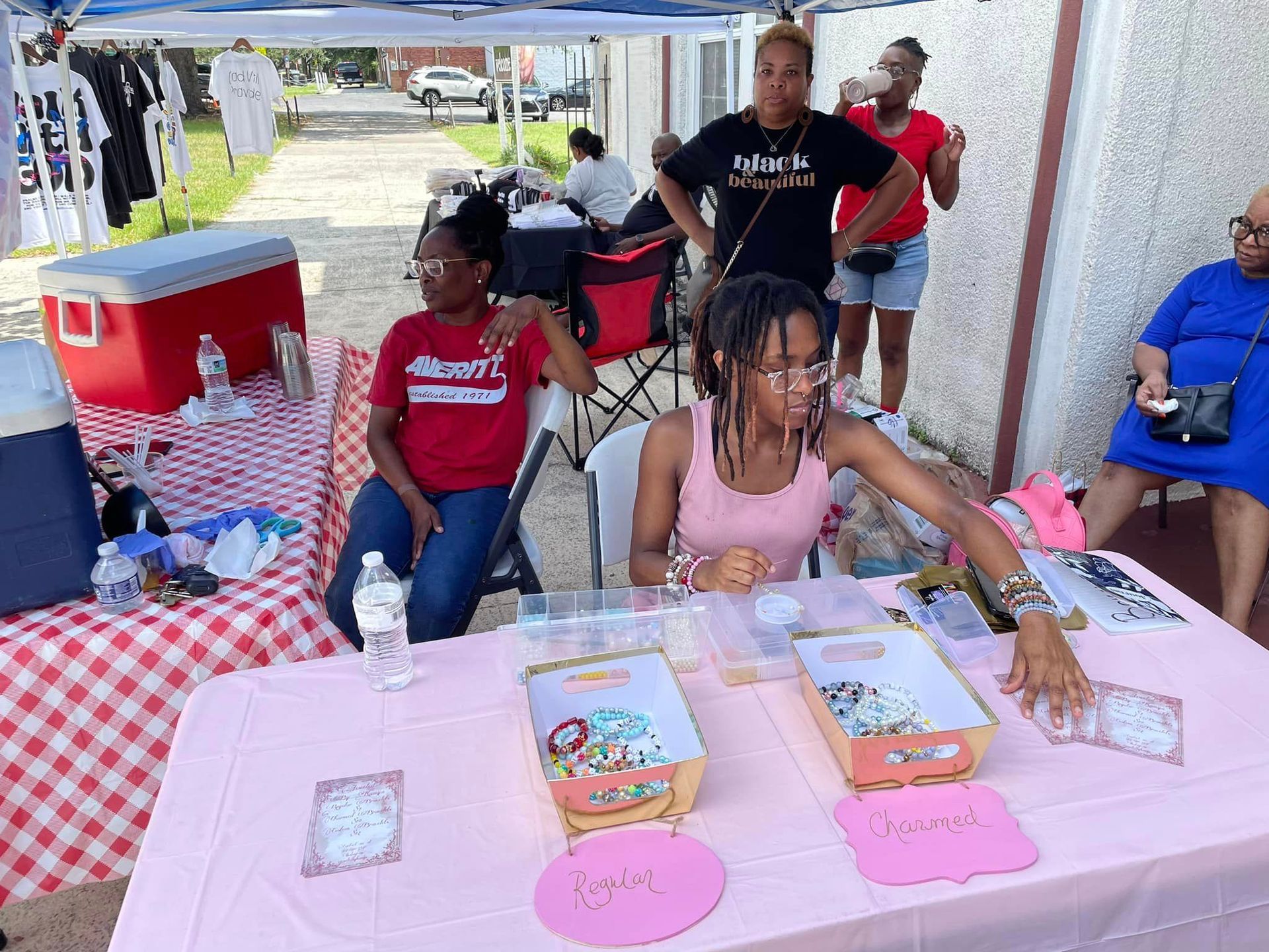 People at a table selling items. Two girls are handling items. Others watch. Pink table cover.