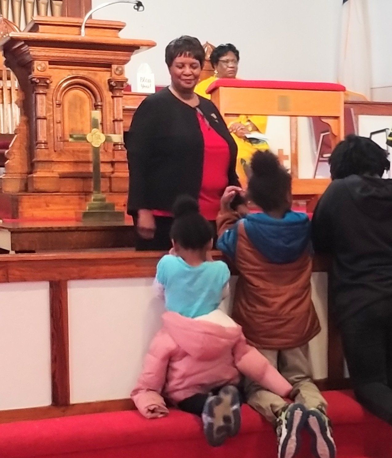 Woman stands at a church altar with children kneeling in front, filming.