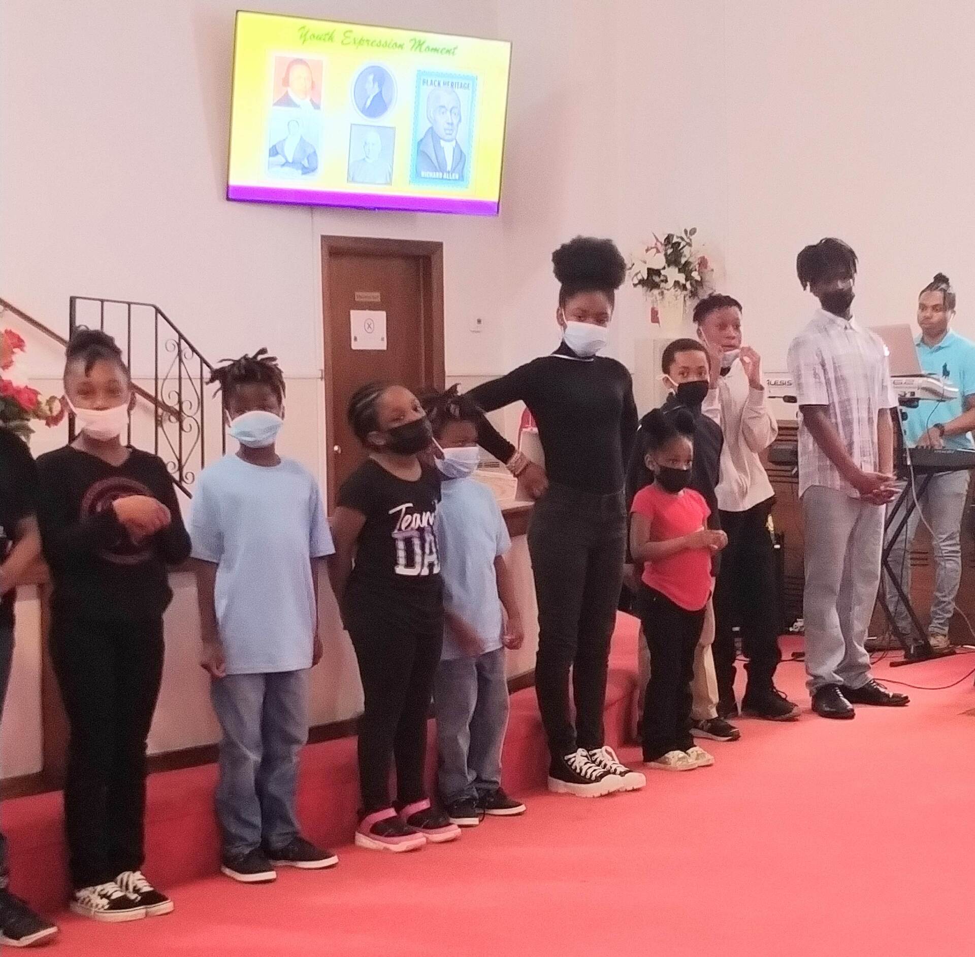 Children standing on stage with a presentation about famous people displayed on a screen.