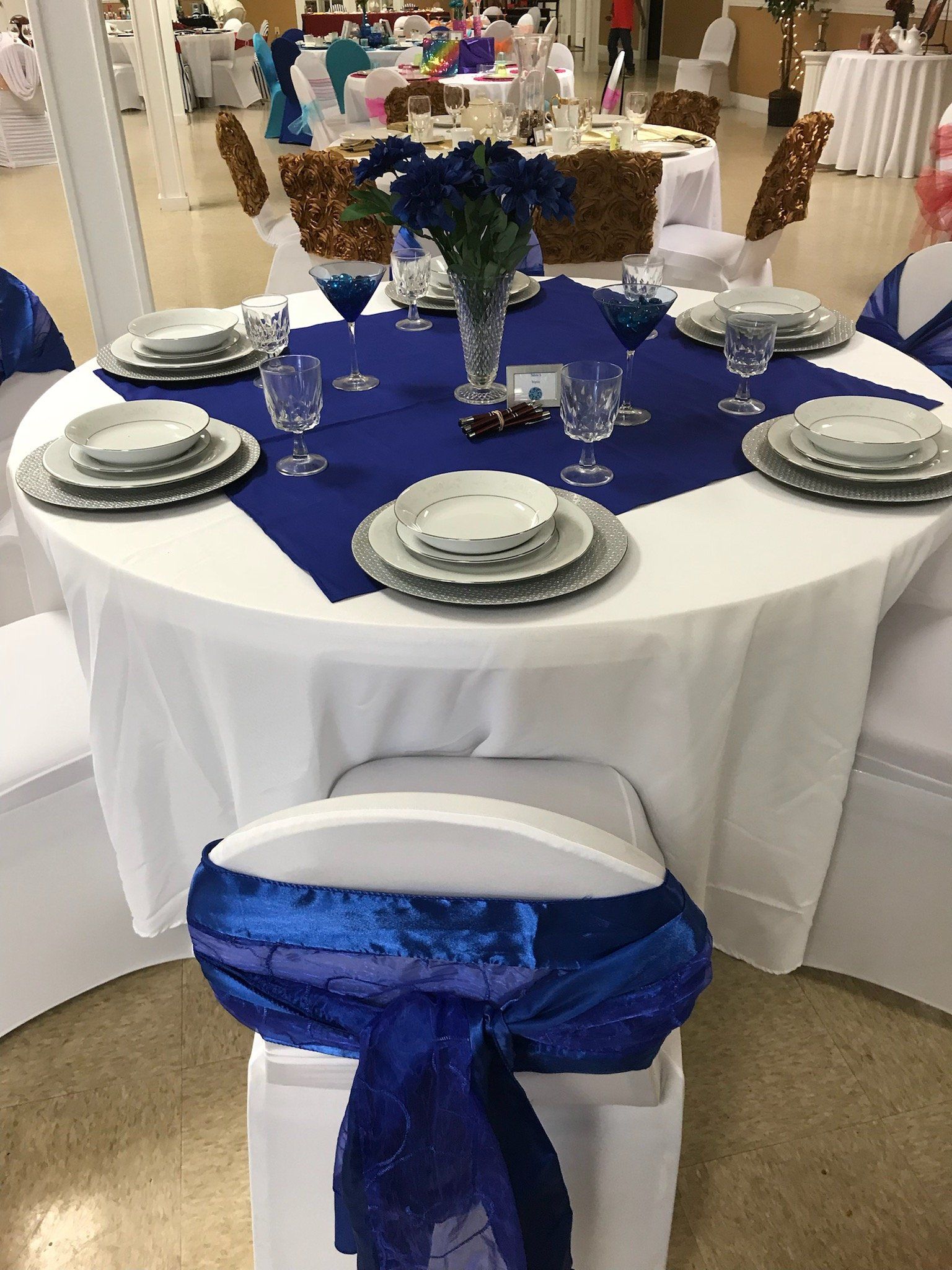 Round table set for a formal event with blue accents: tablecloth, flowers, and chair sashes.