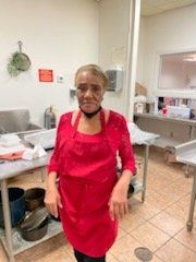 Woman in a red apron stands in a commercial kitchen with pots, sinks, and a window.