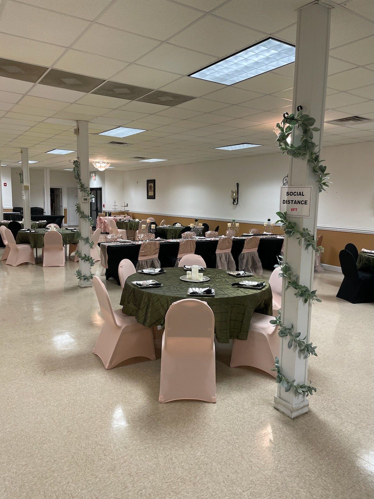Event hall set up with round and rectangular tables, draped in pink, black, and green, decorated with greenery.