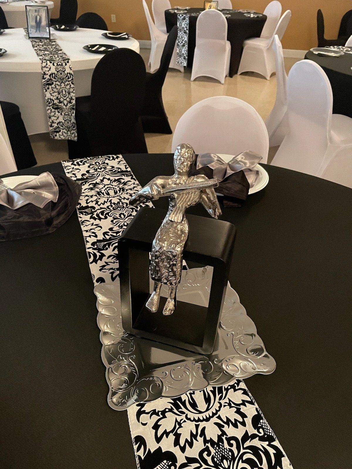 Centerpiece on black table: silver statue on black stand, black and white damask runner, tables with white chair covers.