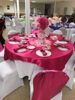 Round table set for a tea party with bright pink tablecloth, napkins, and floral centerpiece, in a room with other tables.