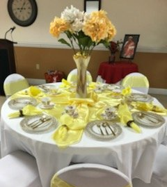 Table set for an event with yellow and white decor, including flowers, plates, and linens.