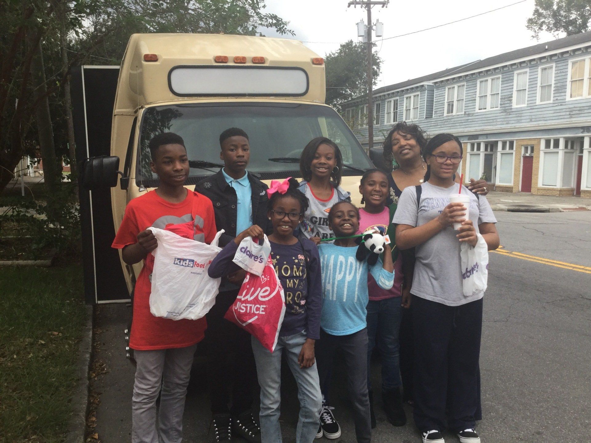 Group of people stand in front of a tan van. Some hold bags, smiling. Buildings in background.