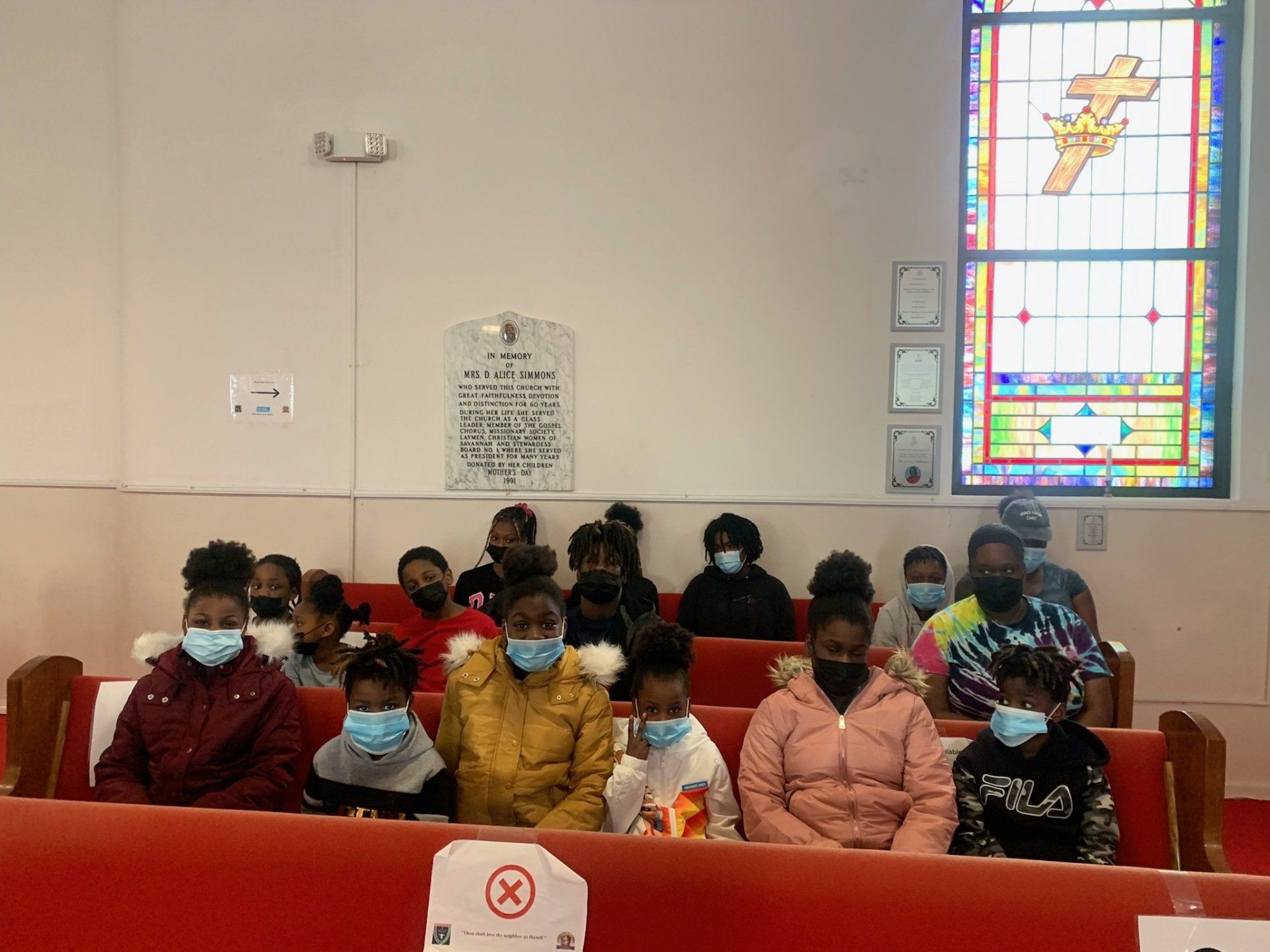 Group of people wearing masks seated in church pews, facing forward, near a stained glass window.