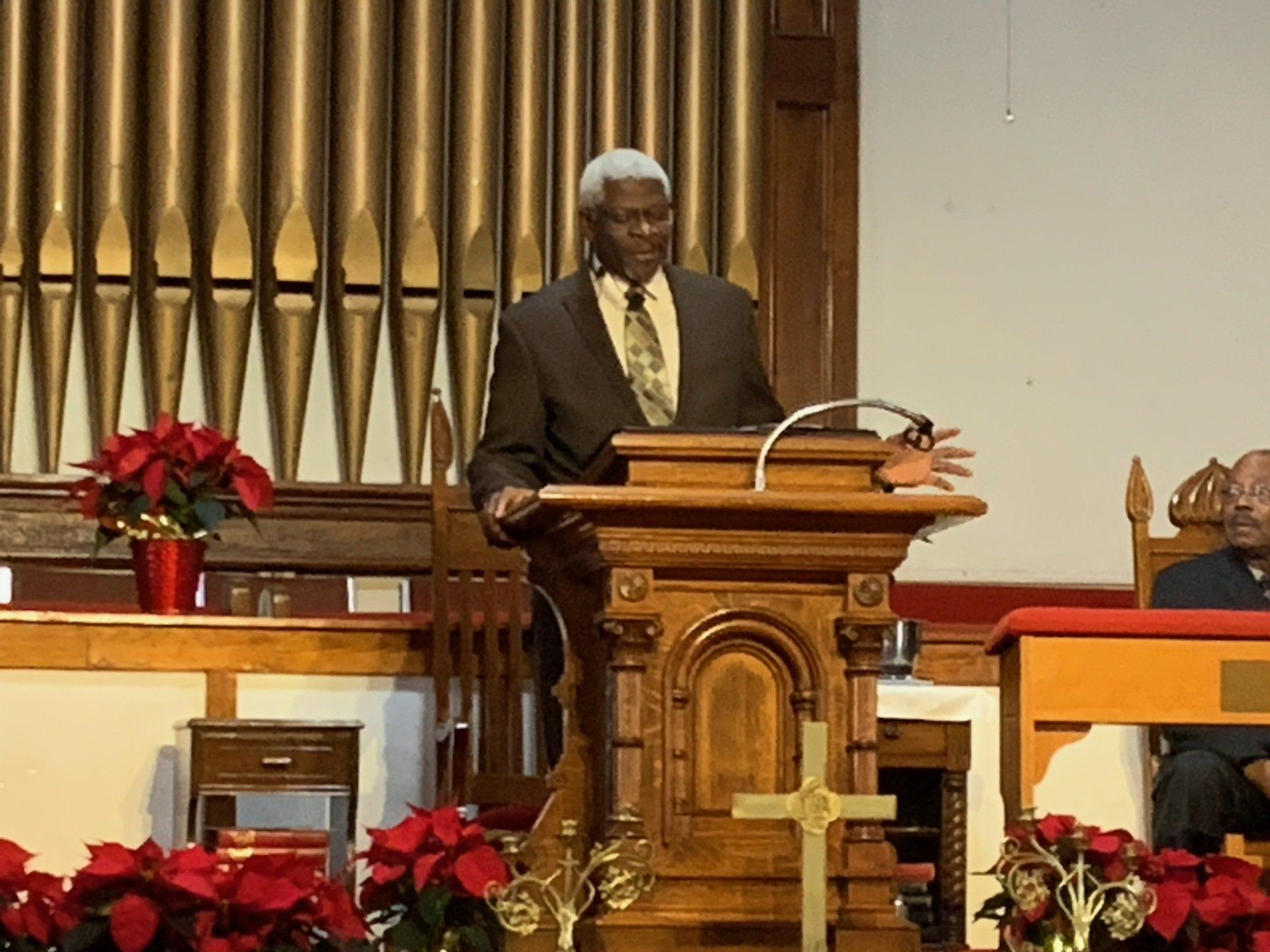 Man speaking at a wooden lectern in a church. Behind him is an organ and poinsettias.