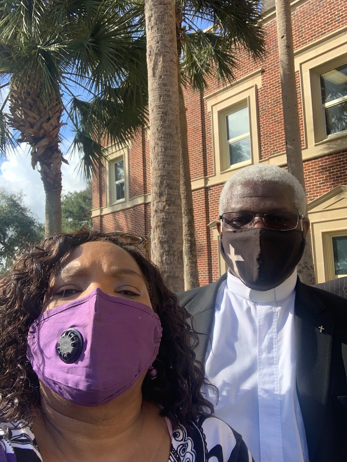 Two people wearing masks pose in front of a brick building and palm trees. One mask is purple, the other black with a cross.