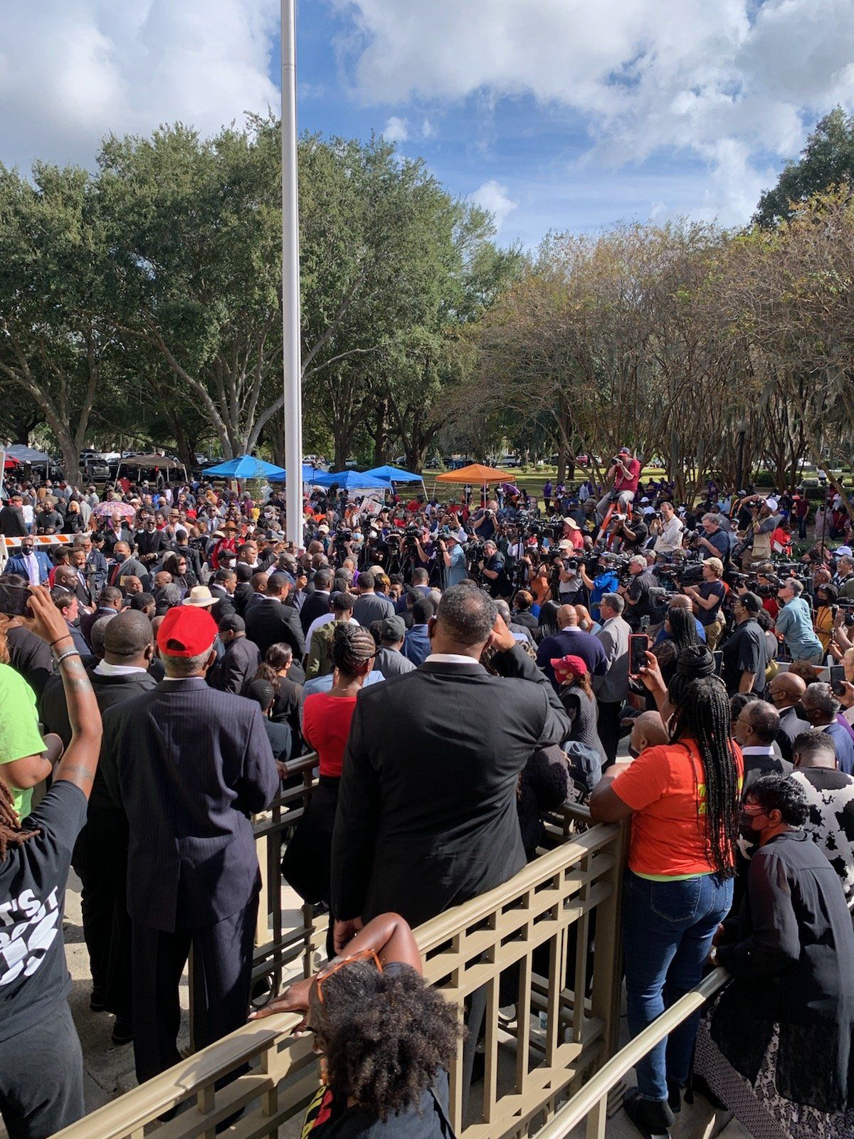 Large crowd gathered outdoors near a building; many people. Cloudy sky. Several people wearing hats.