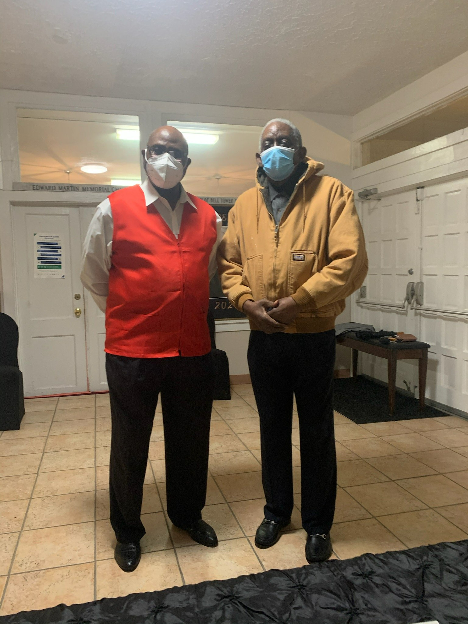 Two men wearing face masks, one in a red vest, other in a tan jacket, standing inside.