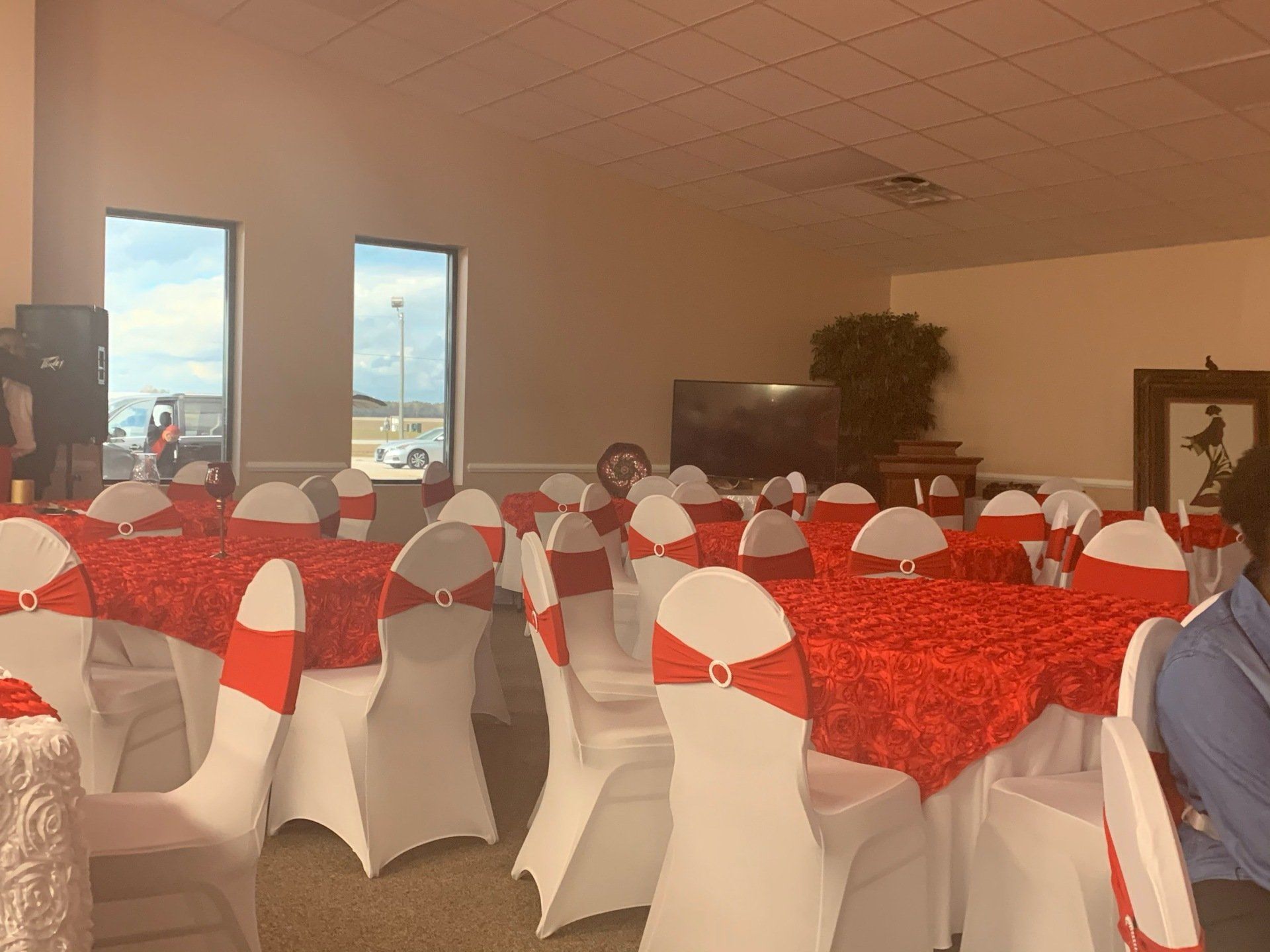 Tables set for an event with red tablecloths and white chairs decorated with red sashes in an interior space.