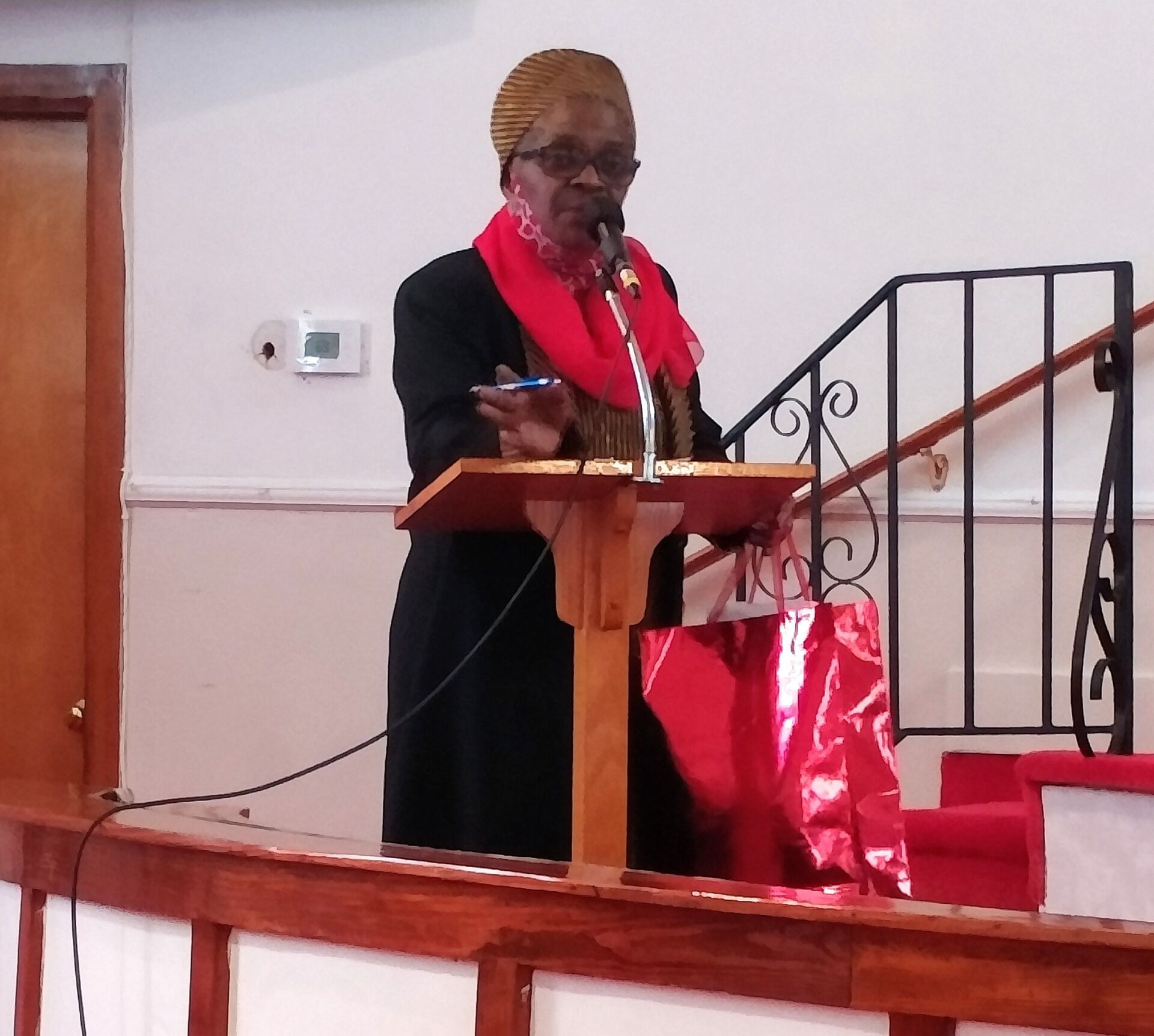 Person speaking at a podium with a microphone, wearing a red scarf in a church.