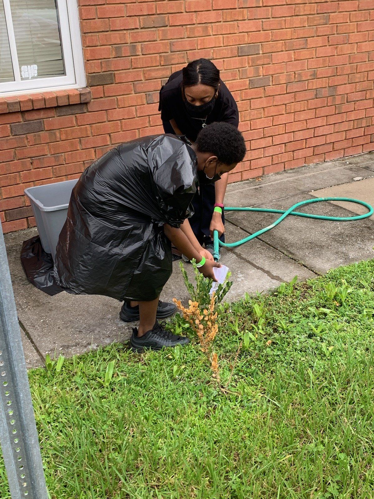 Two people, one in a trash bag, tend a plant with a hose by a brick building.