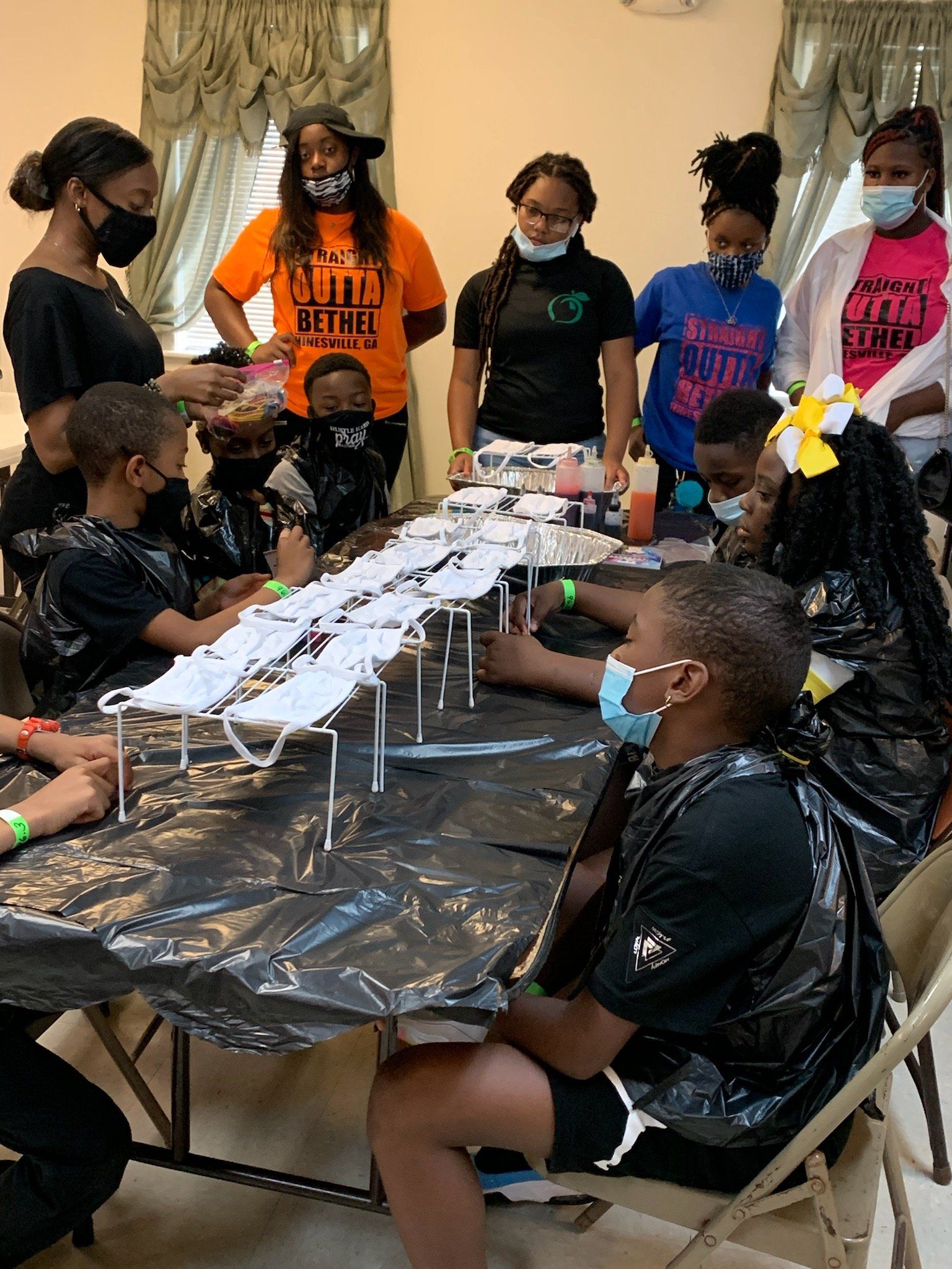 Group of people tie-dyeing shirts at a table. Several adults supervise children.