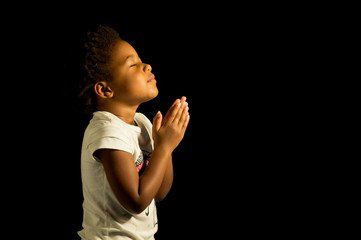 Young person with closed eyes and clasped hands, lit against a black background.