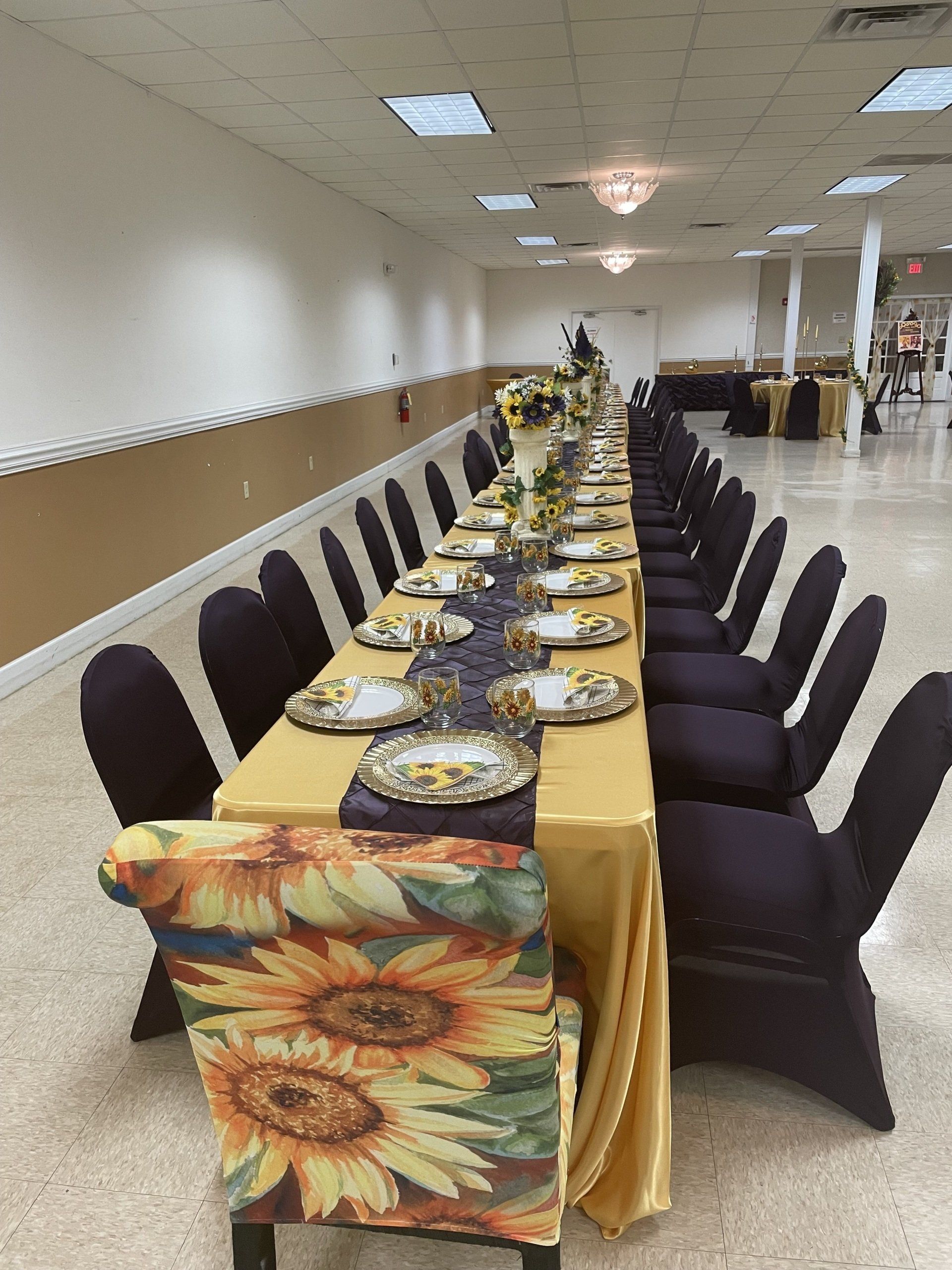 Long table set for a formal event with sunflower-themed chair at the front, gold tablecloth, and purple runner.