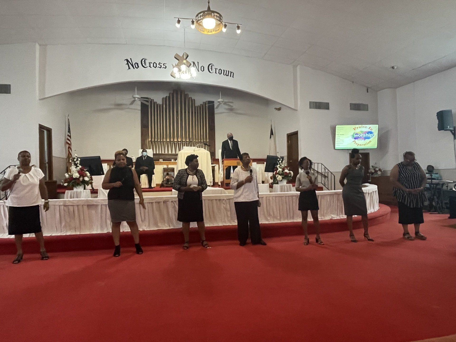 Choir singing on a red-carpeted stage in a church. A preacher is at a podium, and a screen displays a logo.