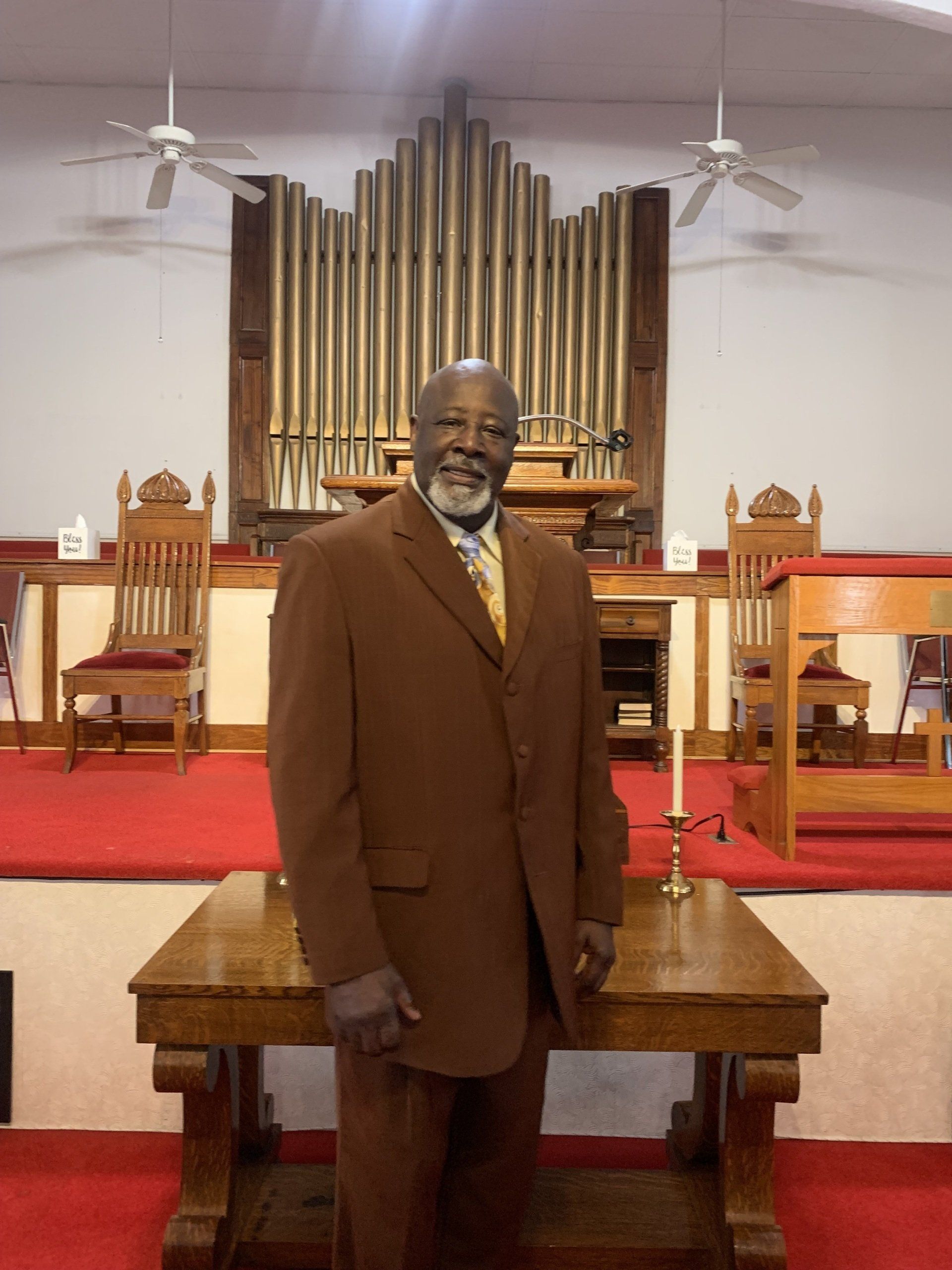 Man in brown suit standing in front of church organ and altar.