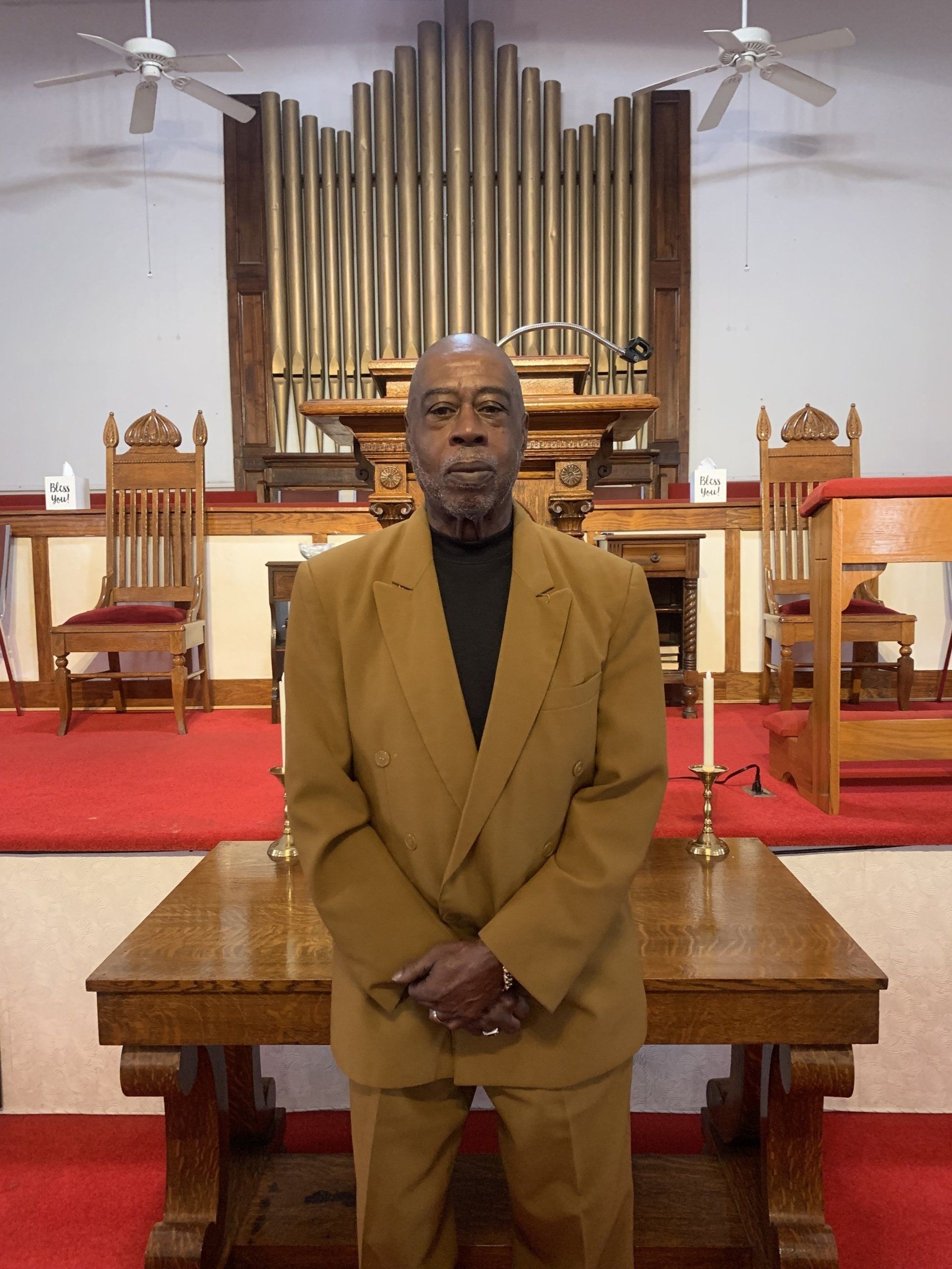 Man in a tan suit standing in front of a church altar with an organ in the background.