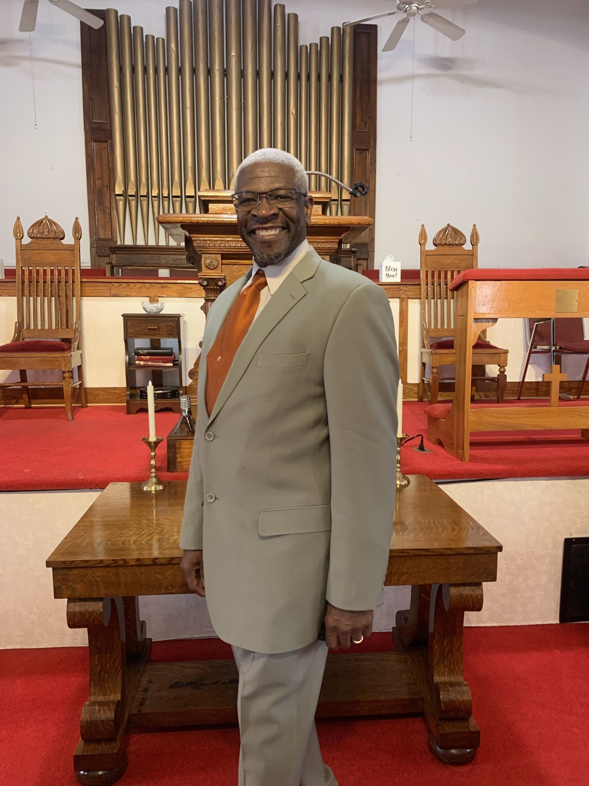 Man in light suit smiles in church, stands before altar, pipe organ in background.
