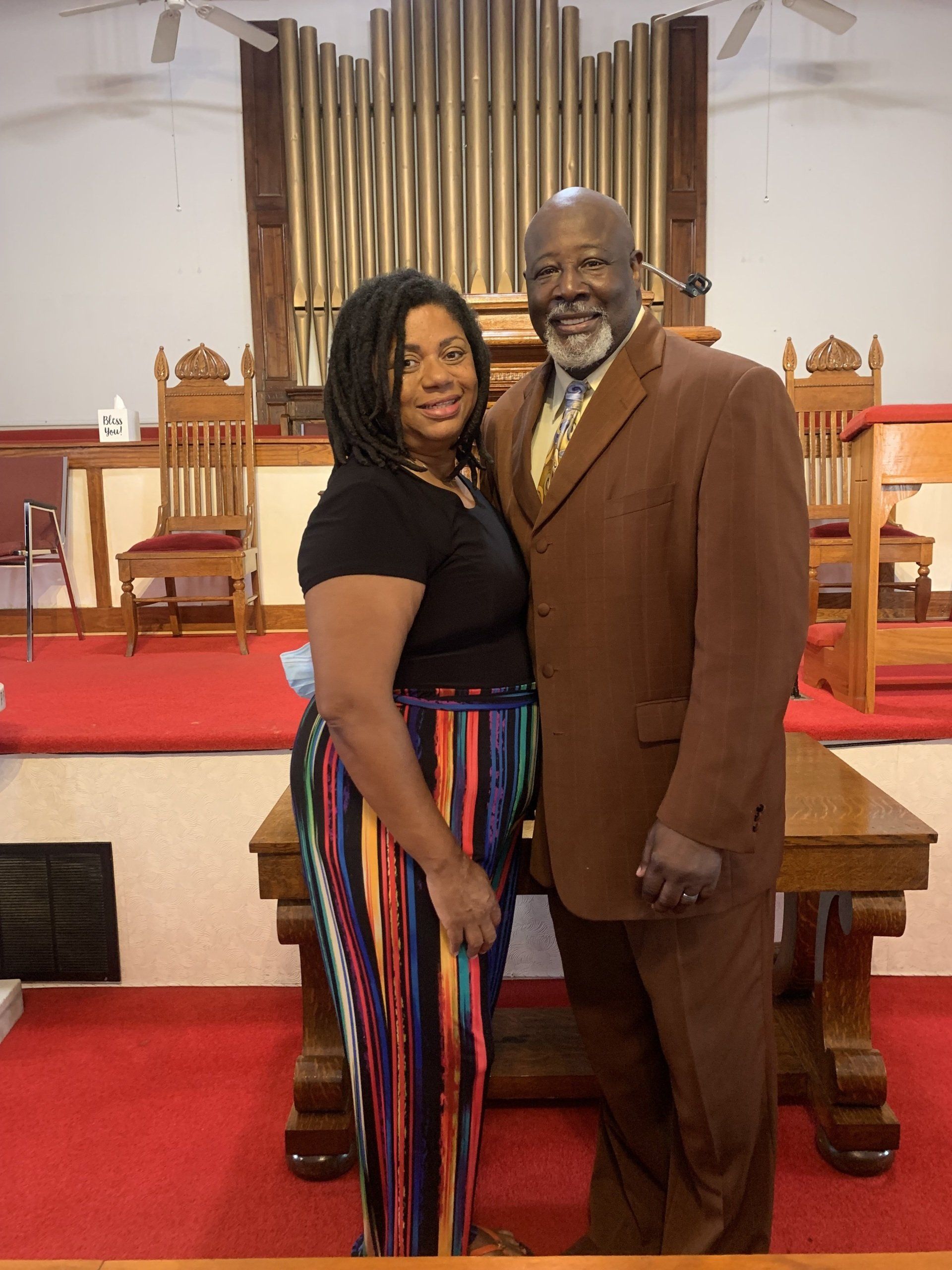 A man and woman stand together in a church. The man wears a brown suit. The woman wears a black top and striped pants.