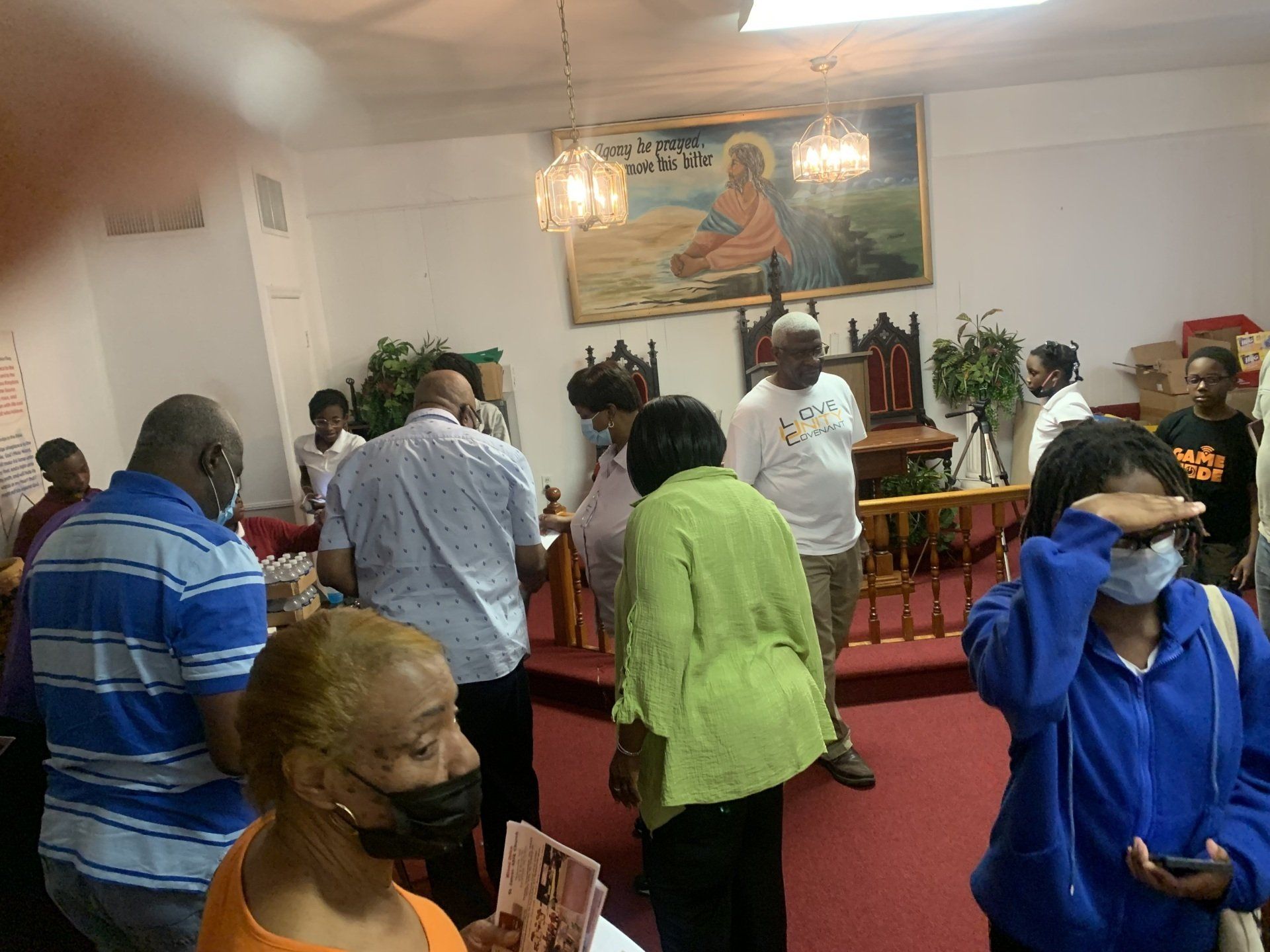 People gathered inside a building with a stage, possibly a church. Some wearing masks. A painting is on the wall.