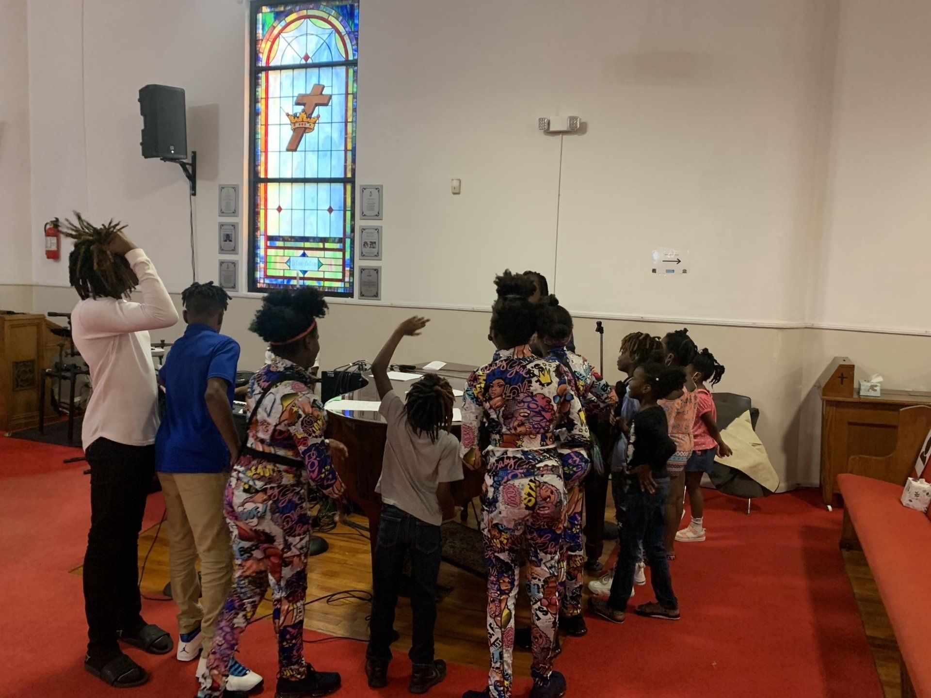 People in a church, near a stained glass window. Some are gesturing, some are looking up. Red carpet.
