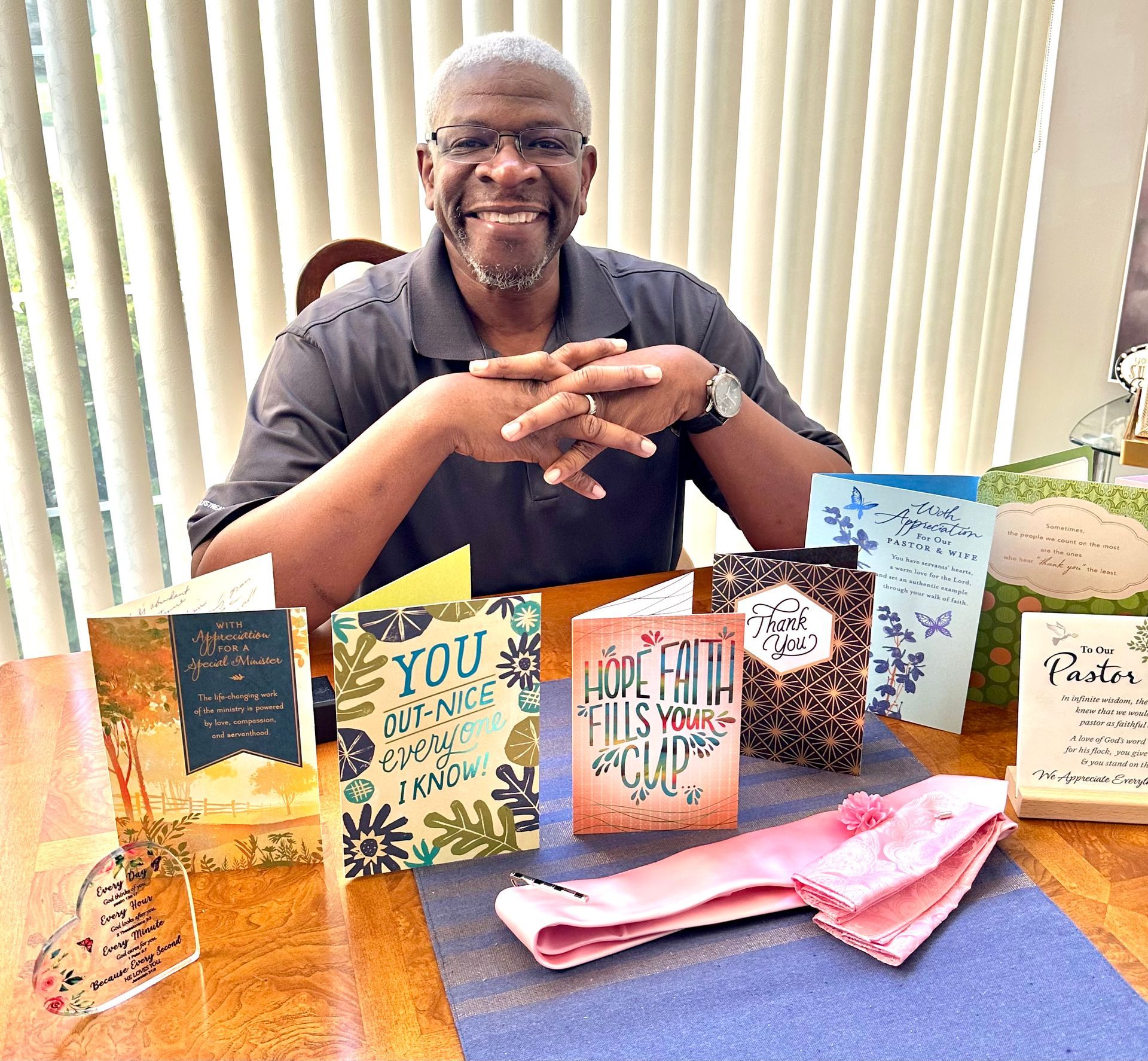 Man smiling, surrounded by cards on a table. Pink handkerchief, blue tablecloth, and window in the background.