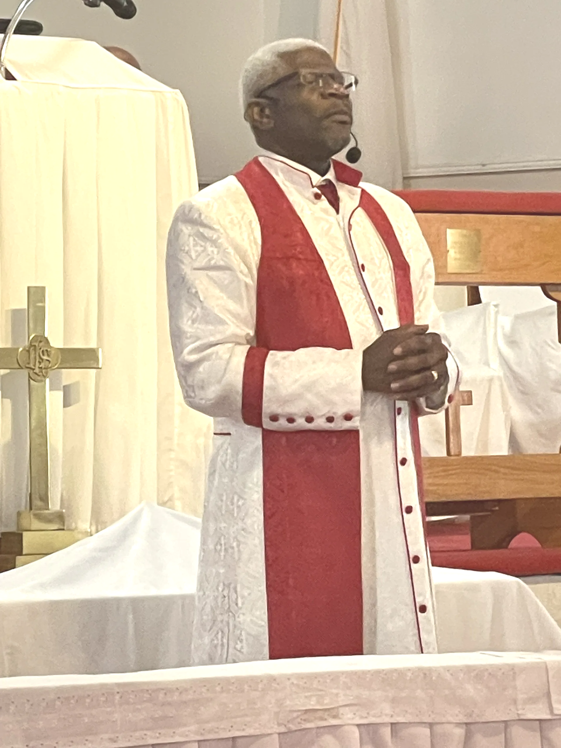 Man in white and red vestments, praying at a church altar, with a cross behind him.