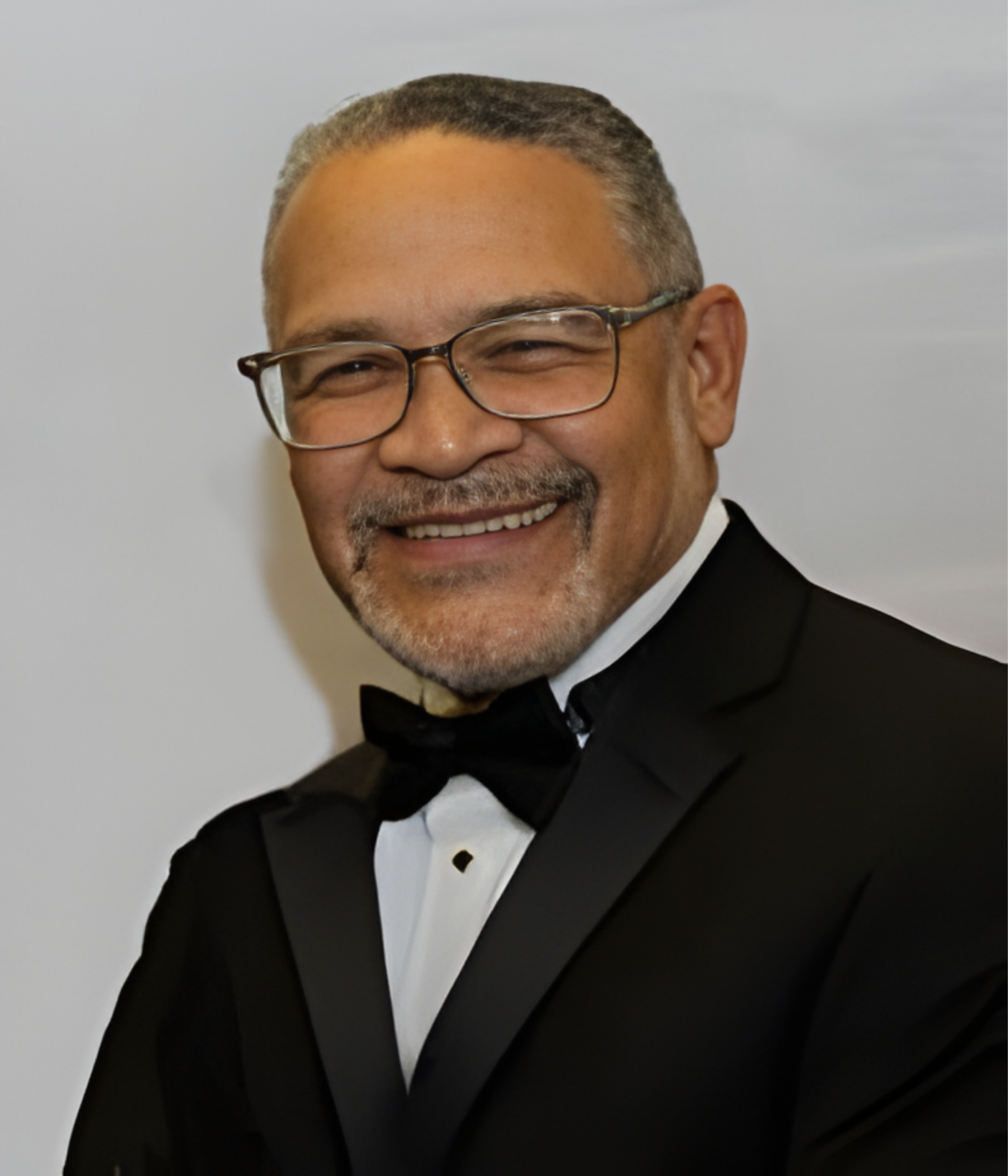 Man in black tuxedo smiling, wearing glasses and a bow tie.