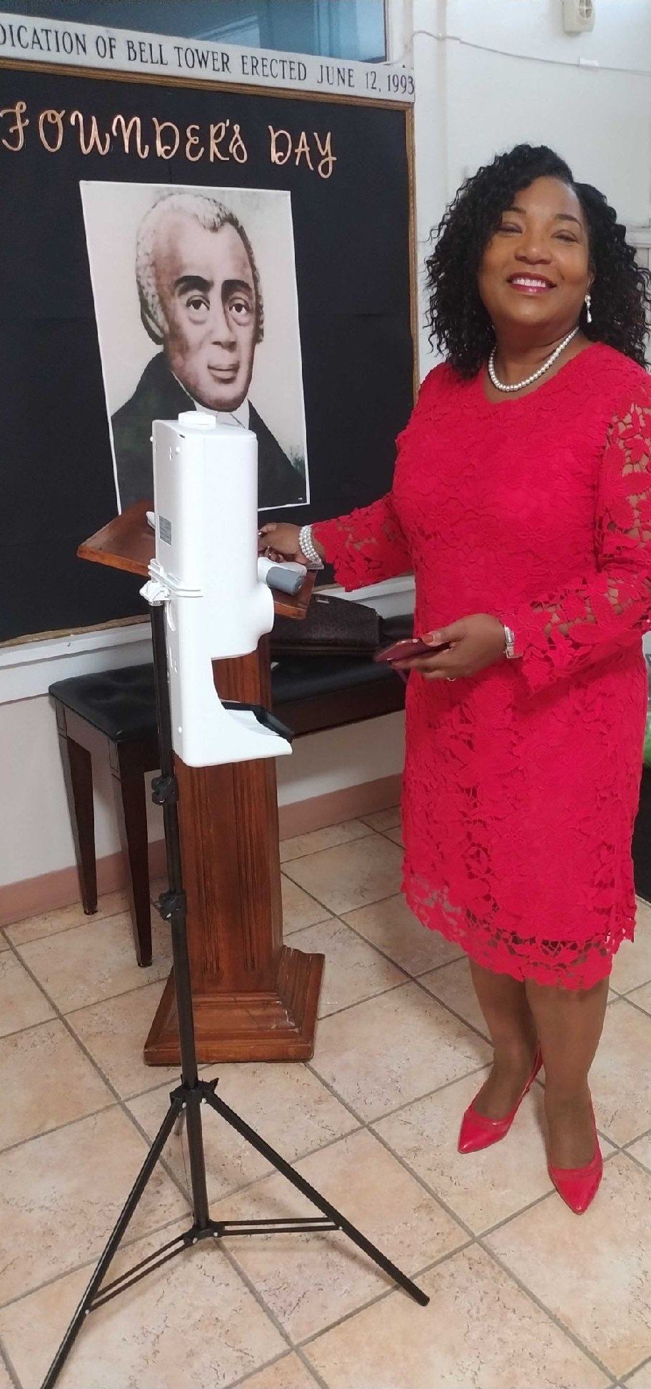 Woman in red dress stands next to a hand sanitizer station, black podium, and Founder's Day display.