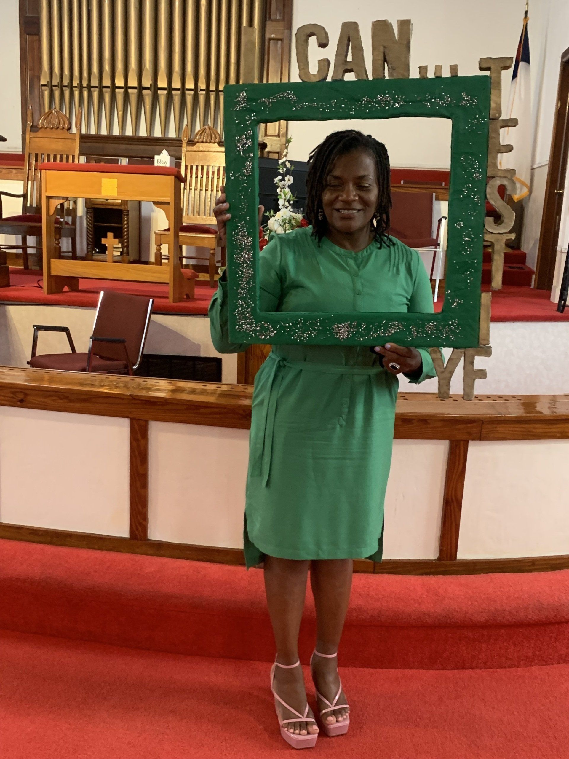 Woman holding a green frame, smiling, wearing a green dress and heels, in a church.