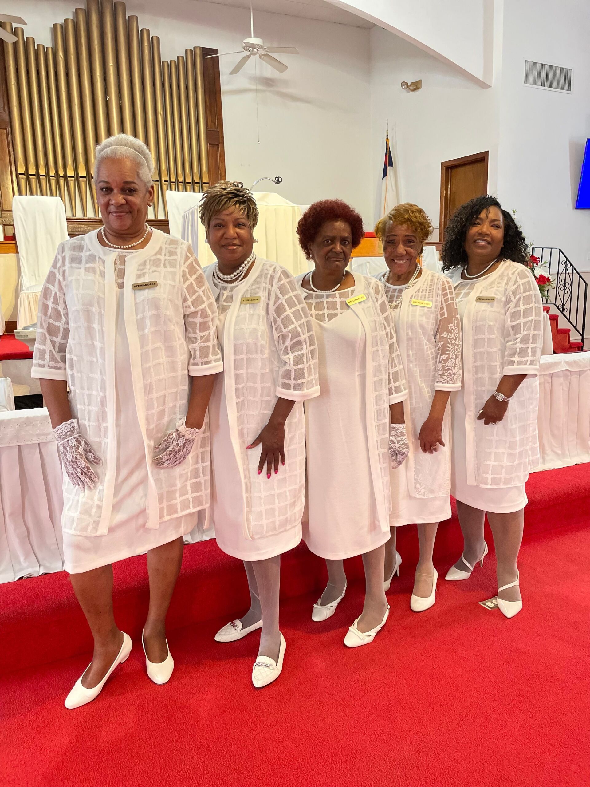 Five women in white dresses and jackets, smiling, standing on a red carpet in a church.