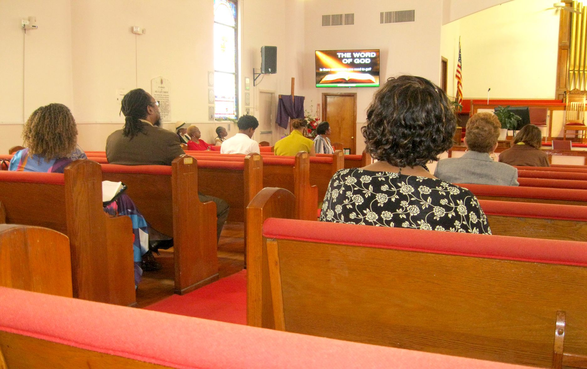 People raising hands in a church, focused on a man in a blue suit. Cross and screen in the background.