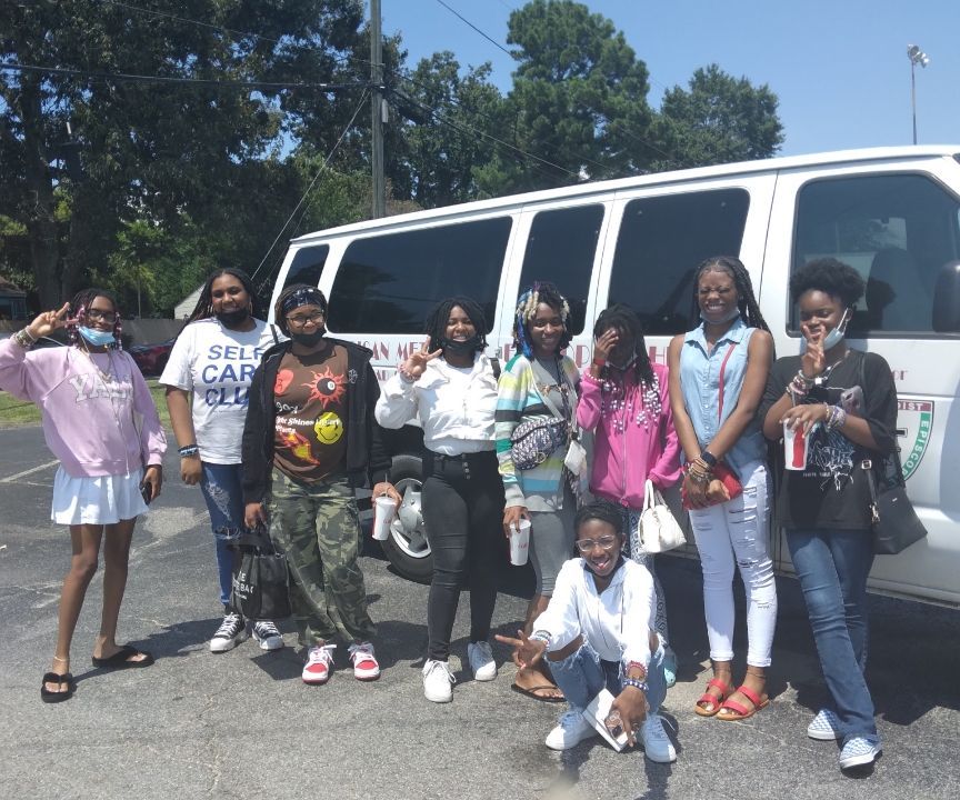 Group of people posing by a van outdoors. Some are holding drinks, making peace signs, and wearing masks.