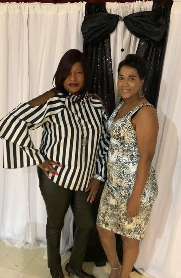 Two women posing before sequined backdrop, one in striped top and pants, the other in silver patterned dress