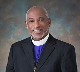 Man in suit with clerical collar smiling; blue and gray background.