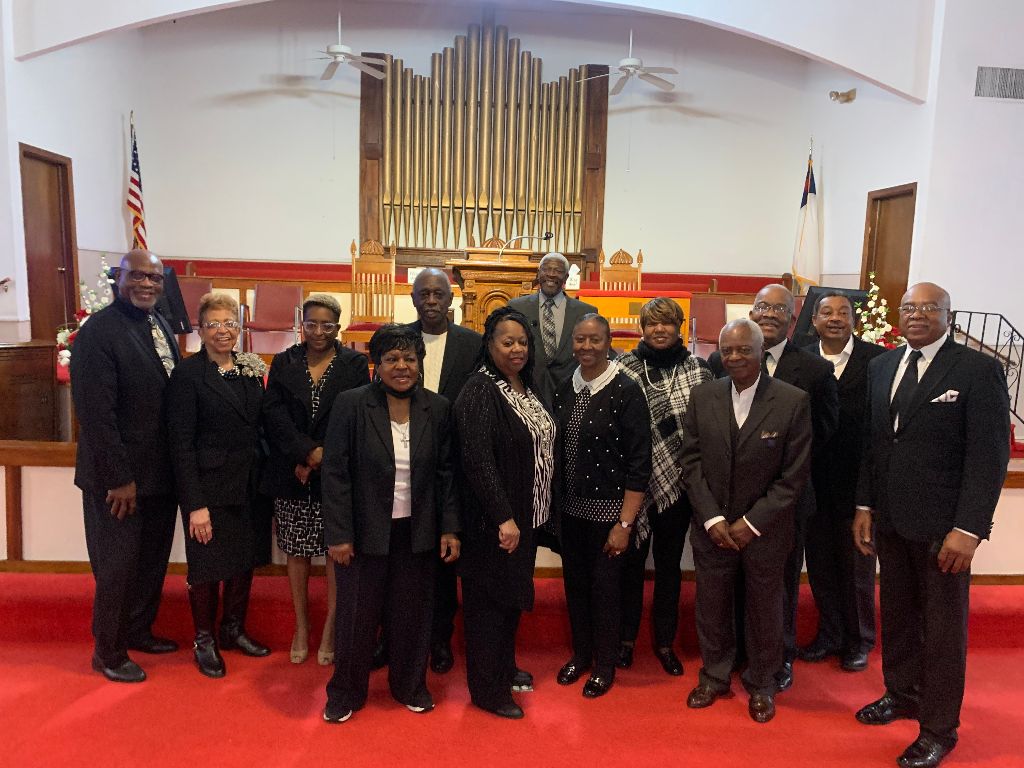 Group of people standing on a red carpet in front of a church organ. They are wearing formal attire.