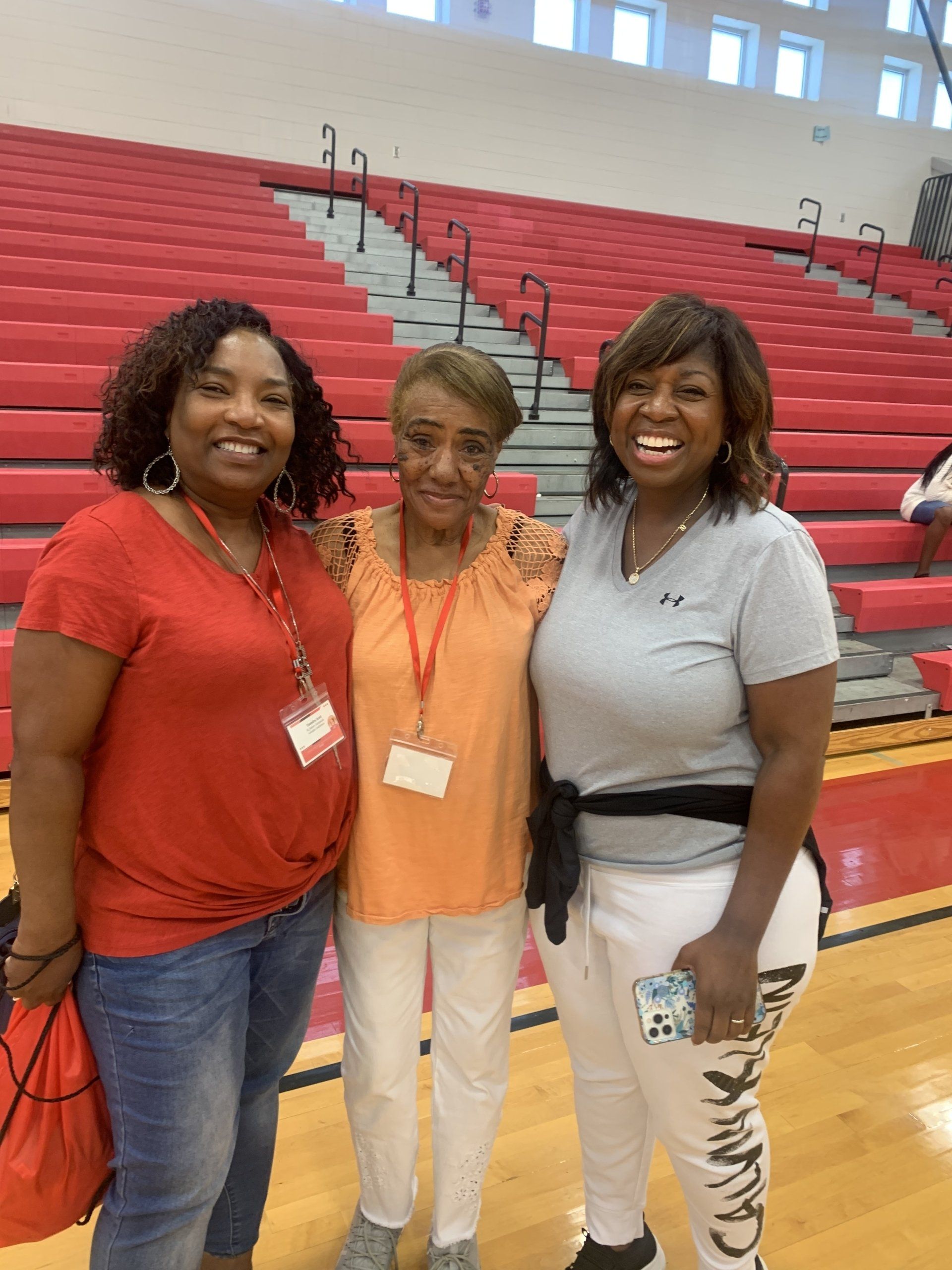 Three women pose together in a gymnasium, smiling. One wears orange, another red, and the third wears gray.