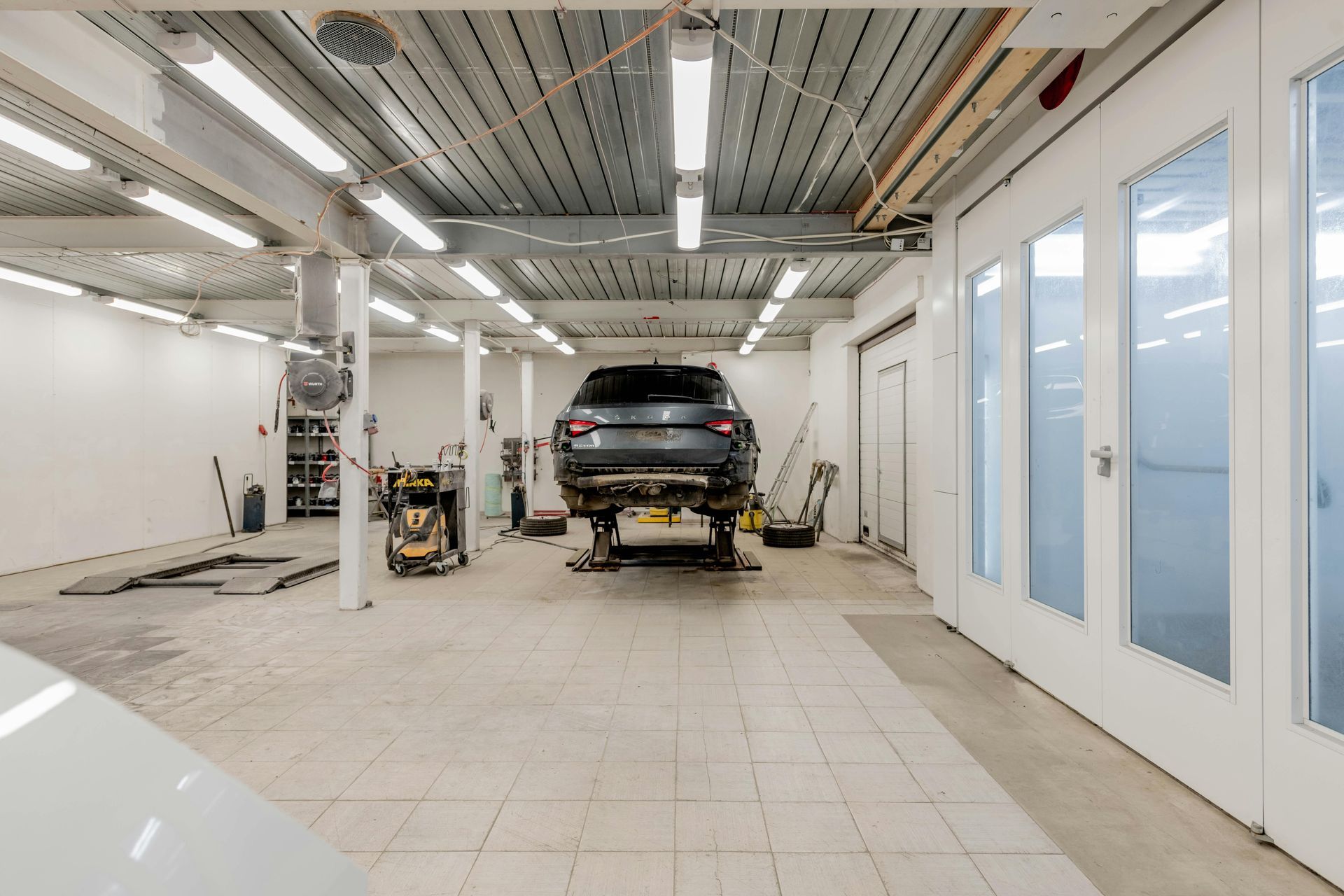 Atelier de réparation automobile avec un SUV noir sur un pont élévateur, sol carrelé blanc et éclairage zénithal intense.