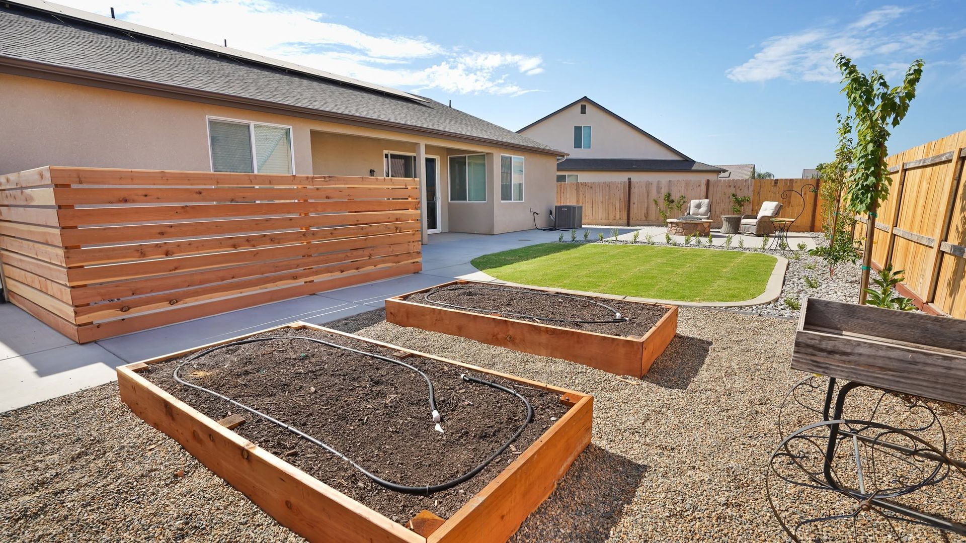 Backyard garden with raised wooden beds, gravel paths, grass, and a house patio under a blue sky