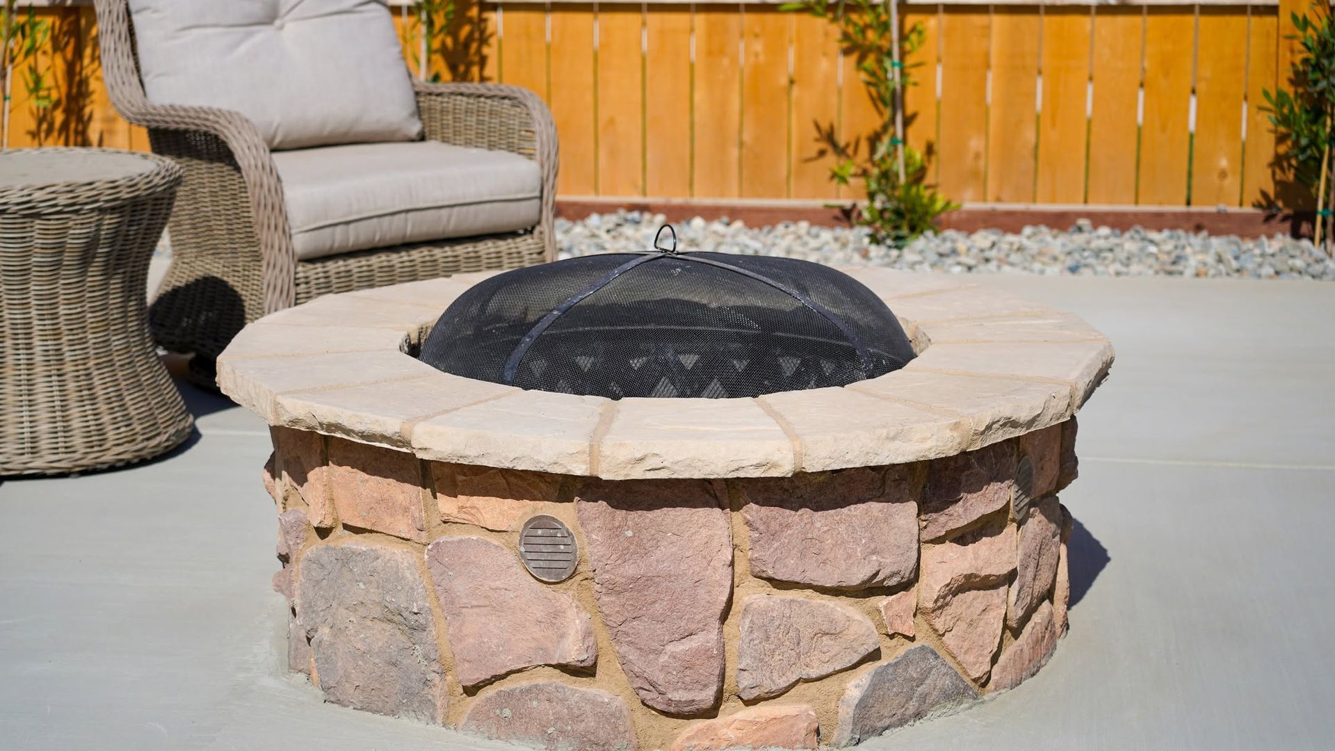 Round stone fire pit on a patio with a wicker chair and wooden fence in the background