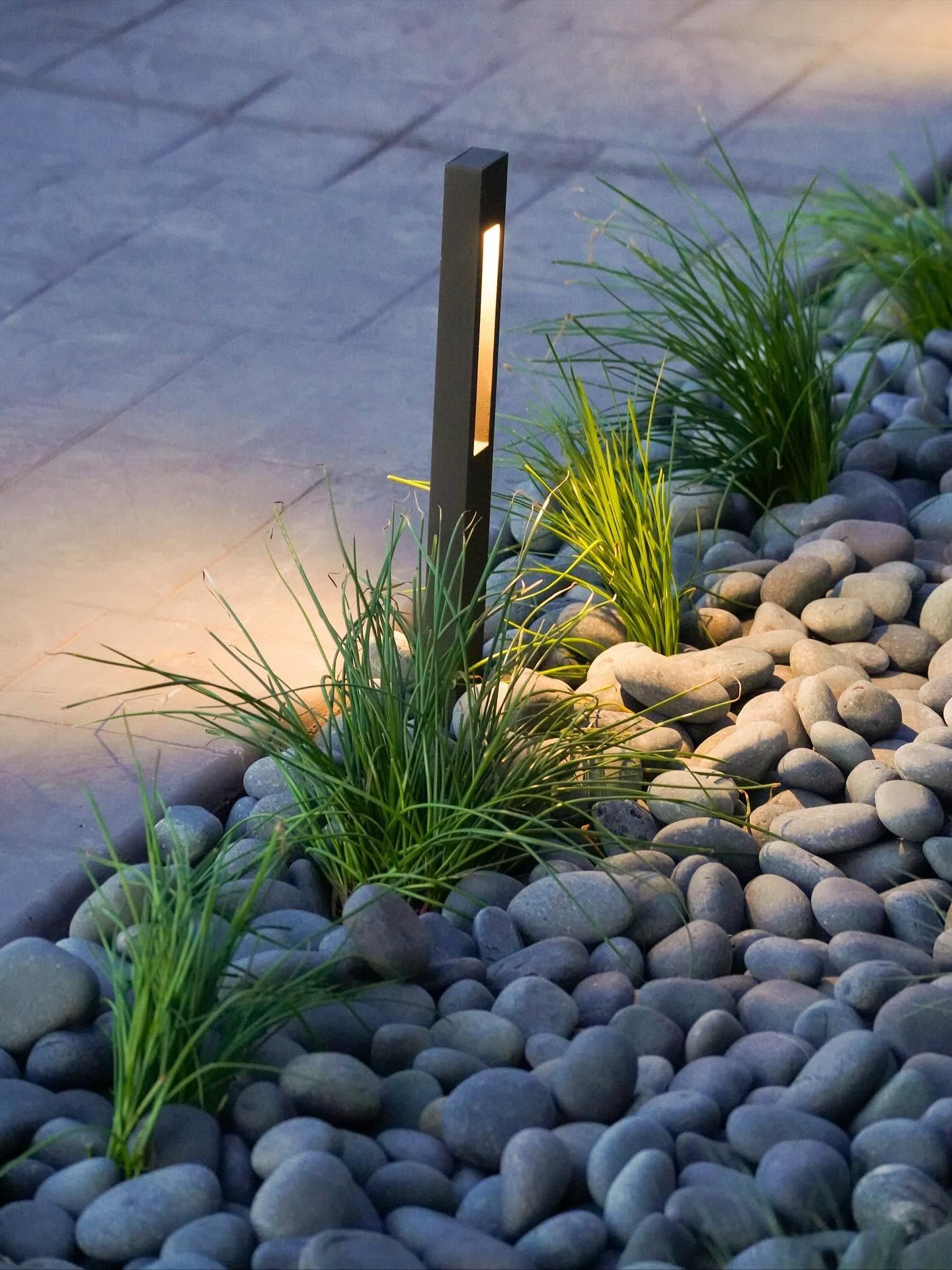 Modern garden path with black pebble stones, grasses, and a glowing pathway light at dusk